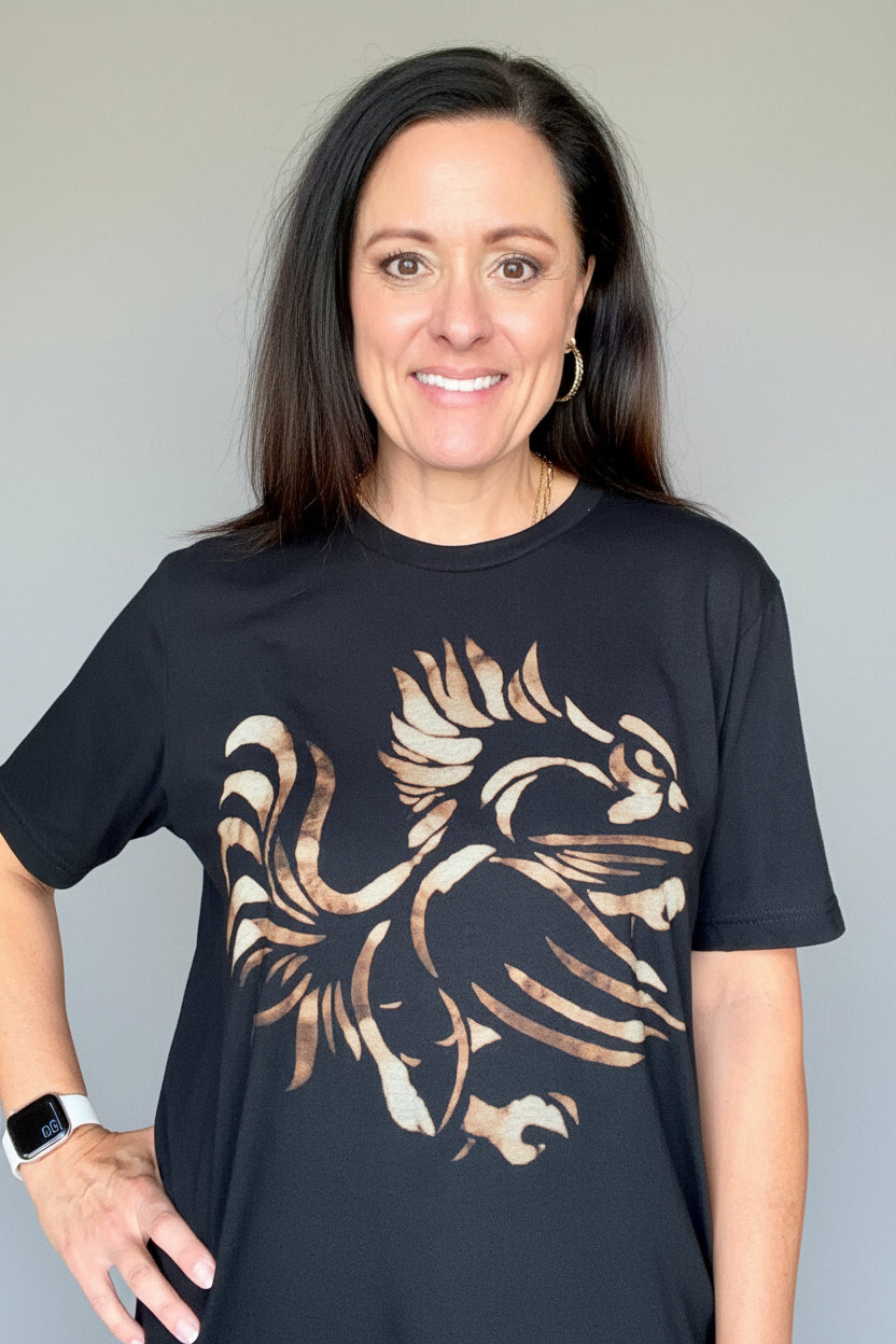A woman wearing a black crewneck t-shirt with a large distressed graphic of a gamecock in a rust color, standing in a home interior hallway.