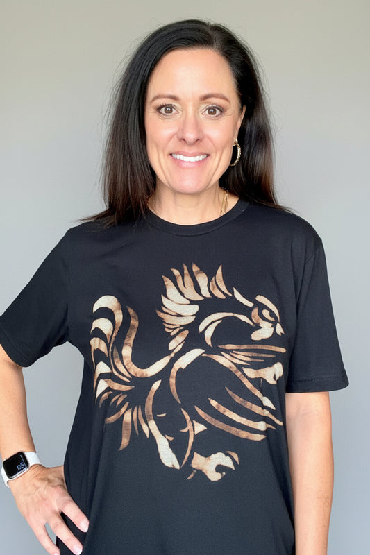 A woman wearing a black crewneck t-shirt with a large distressed graphic of a gamecock in a rust color, standing in a home interior hallway.