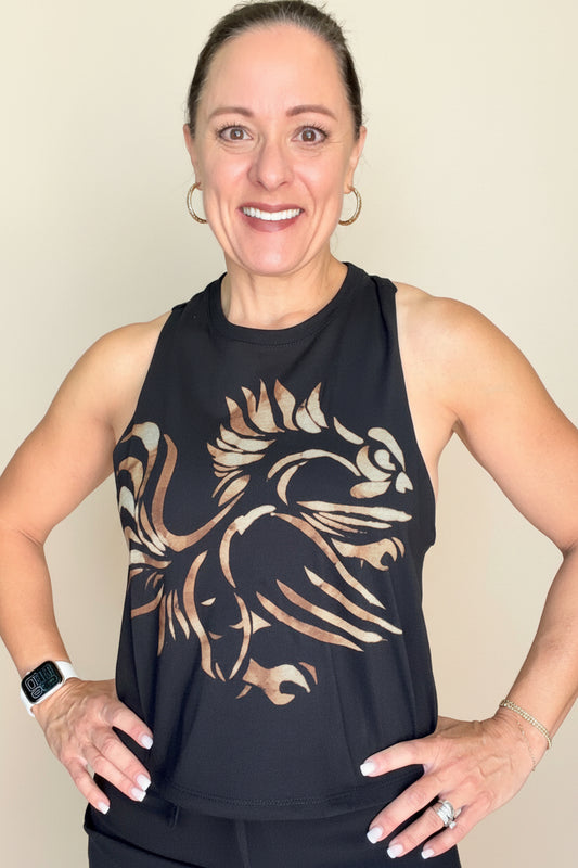 A woman wearing a black sleeveless tank top with a large beige graphic print of a stylized bird, standing in front of an indoor staircase.