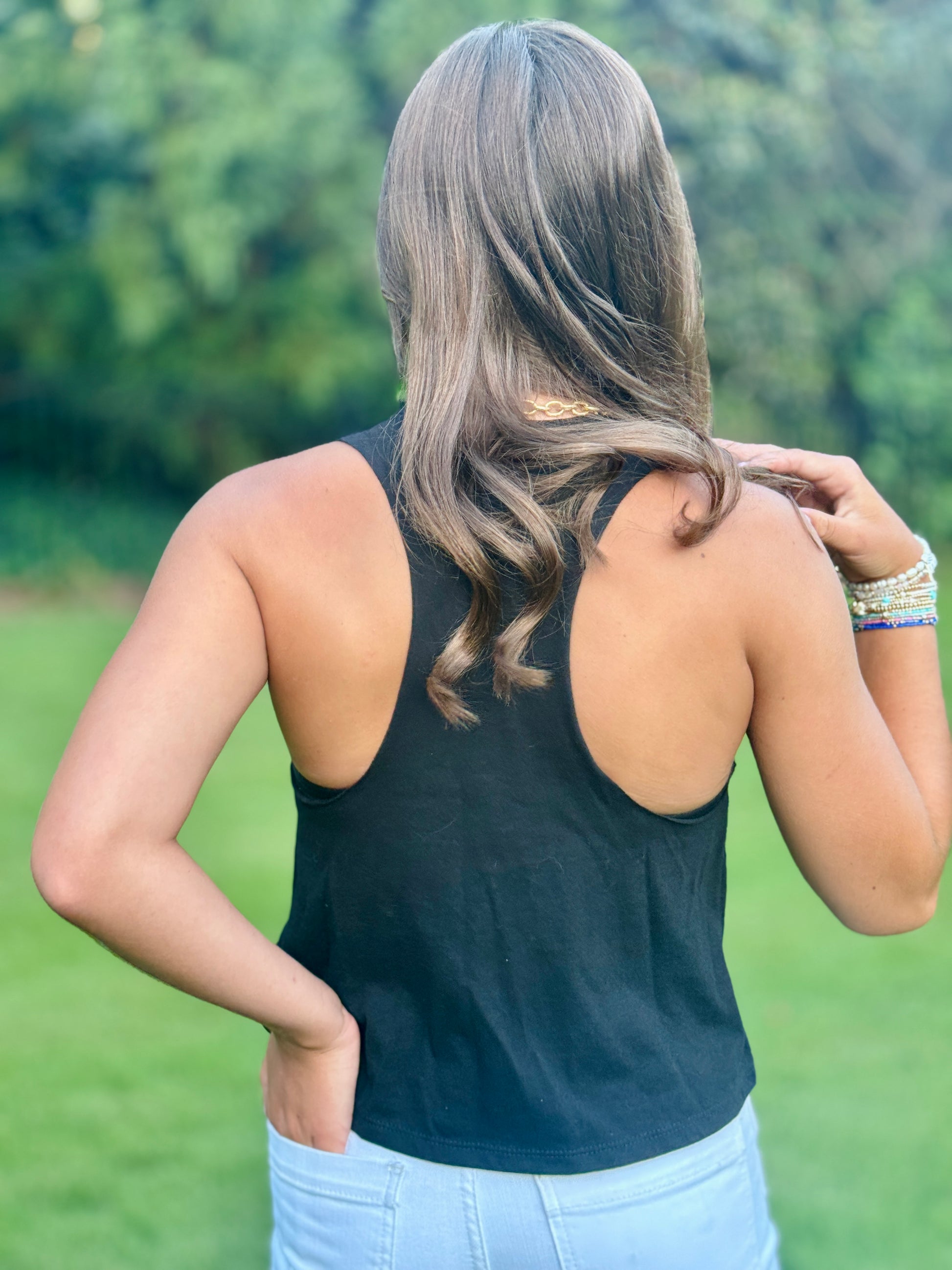 A woman viewed from behind wearing a black racerback tank top and light blue jeans, standing outdoors on a grassy lawn with trees in the background.