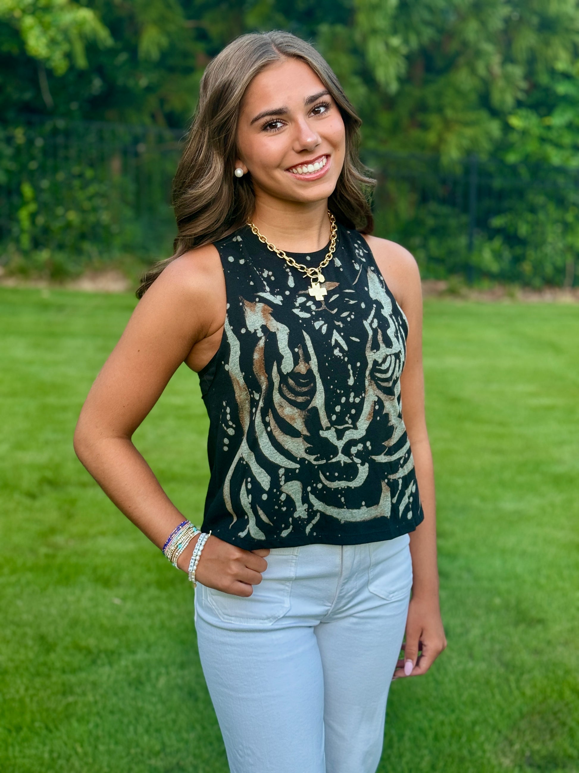 A smiling young woman standing outdoors on a grassy lawn wearing a sleeveless black top with a distressed tiger face print in beige and white, paired with light blue jeans.
